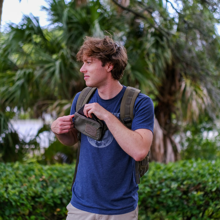 A man wearing a backpack stands outdoors with greenery behind him, opening the GORUCK Sternum Pouch - Cordura.