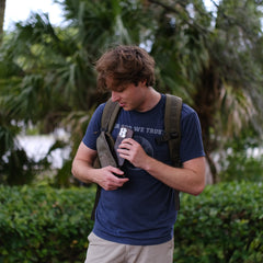 A man with a backpack stands outdoors in front of green foliage, pulling a camera from his Sternum Pouch - Cordura bag.