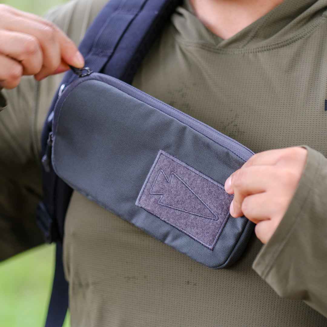 Close-up of a person unzipping the GORUCK Sternum Pouch - Cordura, a small gray pouch attached to their backpack shoulder strap and an essential piece of rucking gear.