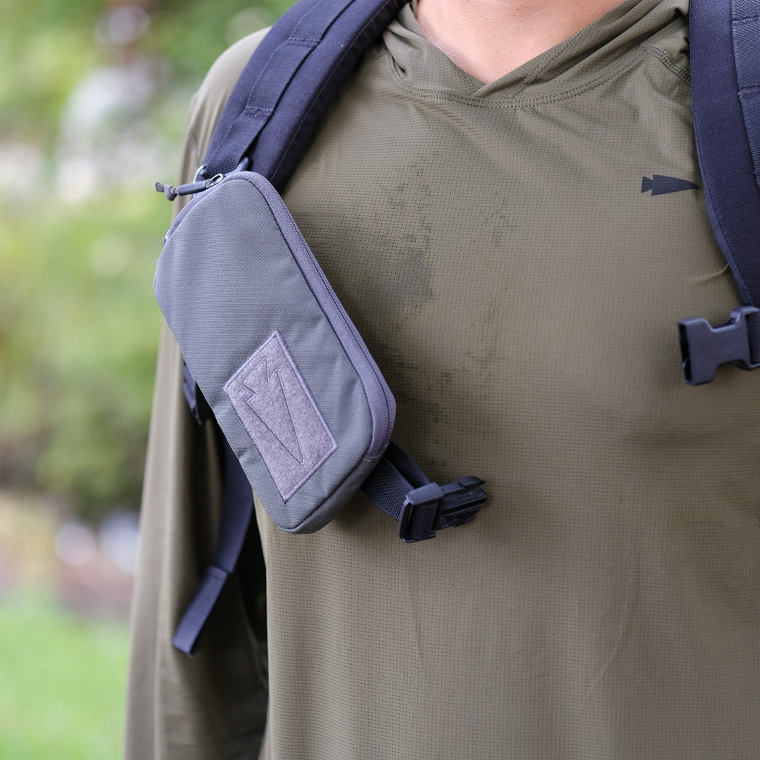 Close-up of a person wearing a green hoodie with the GORUCK Sternum Pouch - Cordura in gray, MOLLE-attached to their black backpack strap—an essential rucking gear accessory.