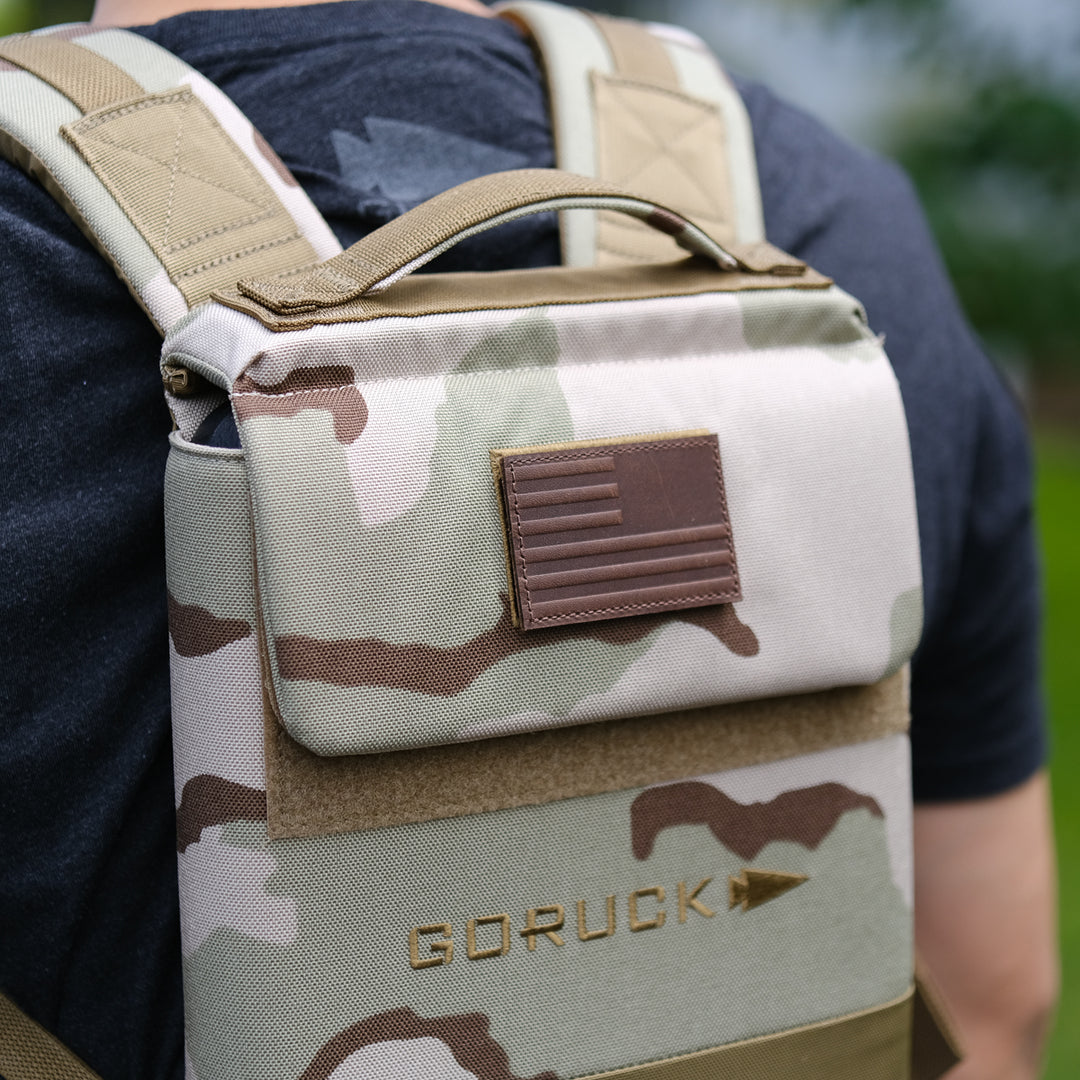 A person wearing a camouflage GORUCK Ruck Plate Carrier 3.0 with a brown American flag patch, designed with ergonomic lumbar support for comfort.