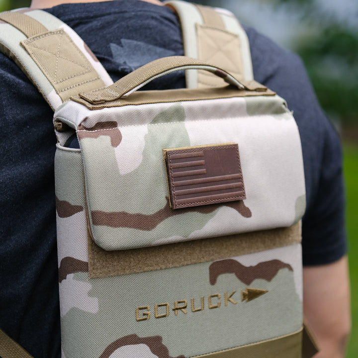 A person wearing a camouflage GORUCK Ruck Plate Carrier 3.0 with a brown American flag patch, designed with ergonomic lumbar support for comfort.