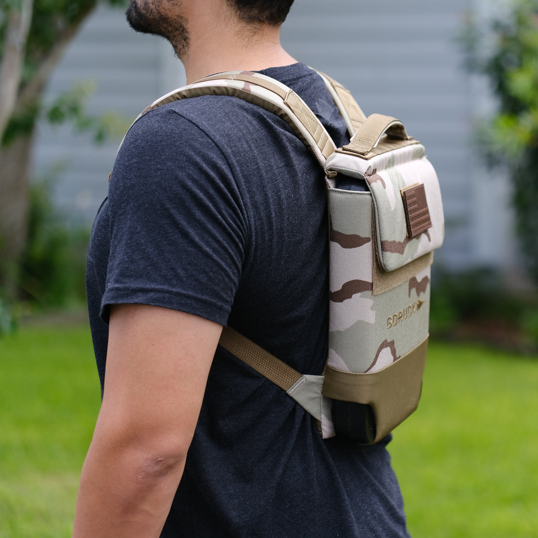 A person stands sideways in a yard wearing the GORUCK Ruck Plate Carrier 3.0, a tan and camo hydration backpack with ergonomic lumbar support.