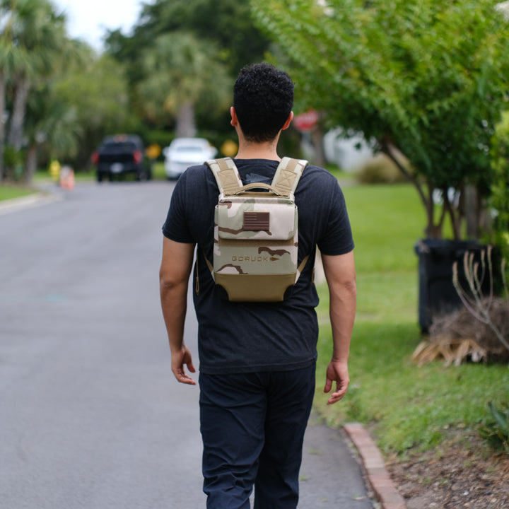 A man walks down a suburban street wearing the GORUCK Ruck Plate Carrier 3.0 backpack, featuring ergonomic lumbar support, paired with dark clothing.