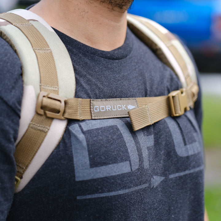 A person outdoors wears a GORUCK Ruck Plate Carrier 3.0 backpack with tan straps over a dark t-shirt, designed for ergonomic lumbar support.