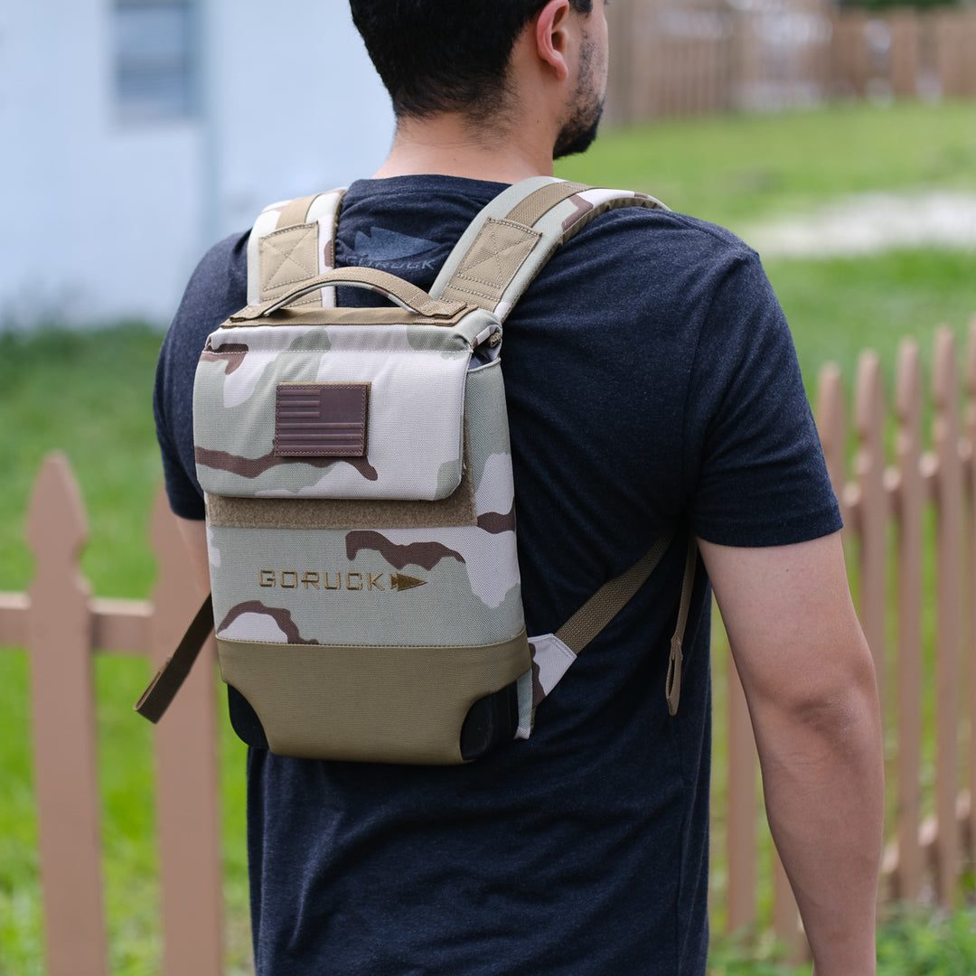A man stands outdoors by a wooden fence wearing the GORUCK Ruck Plate Carrier 3.0, featuring ergonomic lumbar support and a camo pattern.