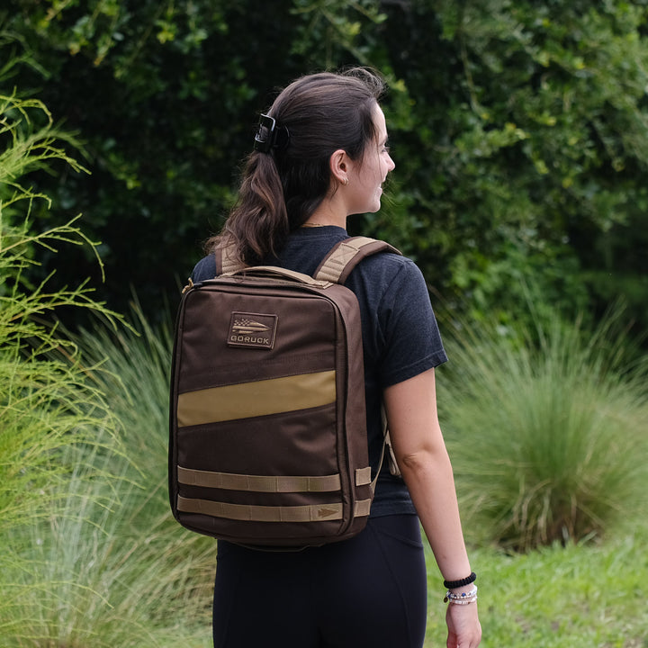 A woman stands outdoors with her back to the camera, surrounded by green plants and grass, wearing a brown GORUCK Rucker 4.0 backpack.