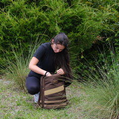 Woman kneeling outdoors with a brown GORUCK rucksack, surrounded by green bushes