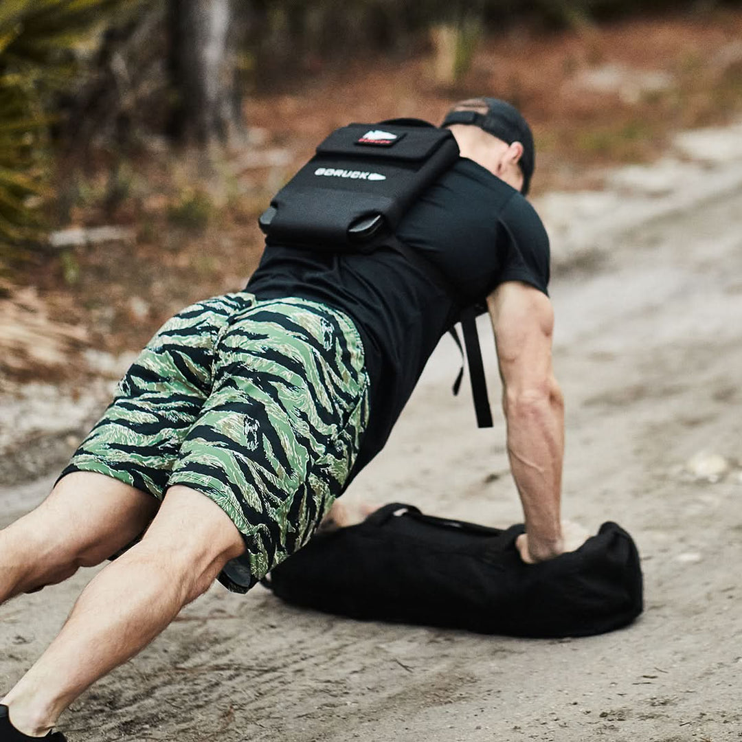 A person in green camo shorts is doing a push-up outdoors on a dirt path, wearing the Ruck Clubs Starter Kit - Light backpack by GORUCK, perfect for rucking.