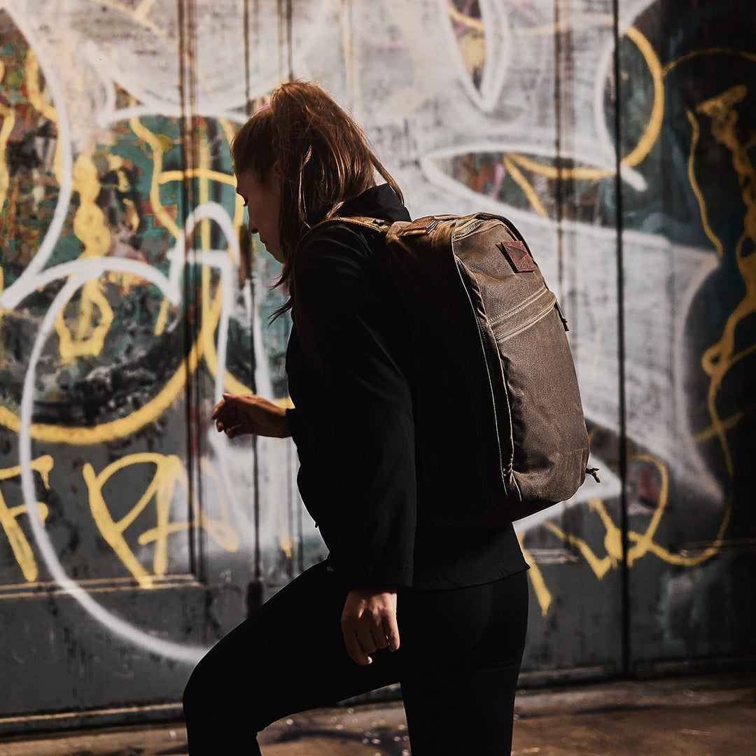 A person with a GORUCK GR1 USA - Heritage backpack stands in front of a wall covered in colorful graffiti.
