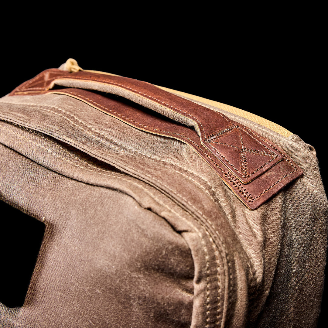 Close-up of the GORUCK GR1 USA - Heritage, a brown waxed canvas rucksack with a leather handle and yellow zipper, set against a black background.