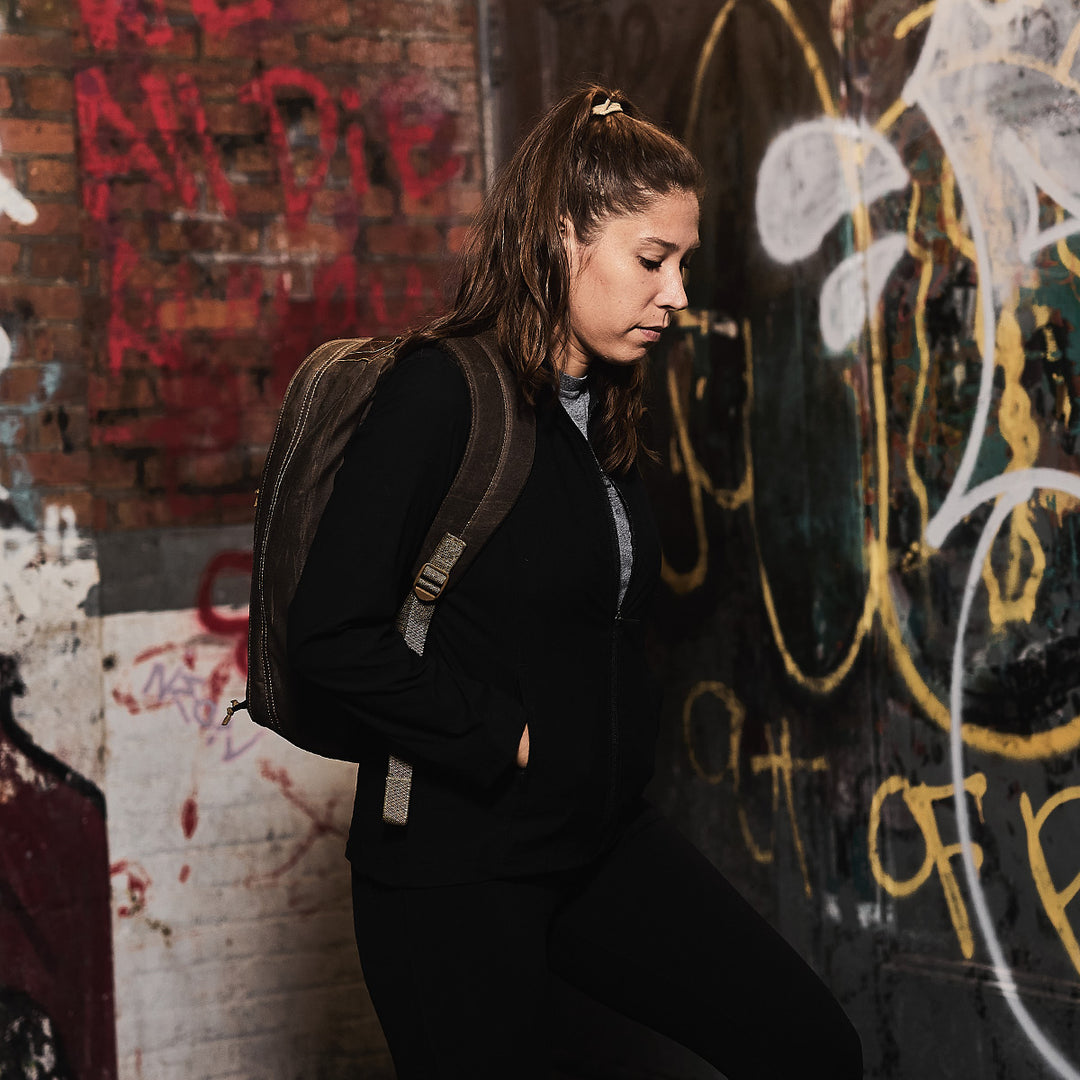 A woman with a GORUCK GR1 USA - Heritage waxed canvas rucksack leans against a graffiti-covered wall, gazing down thoughtfully.