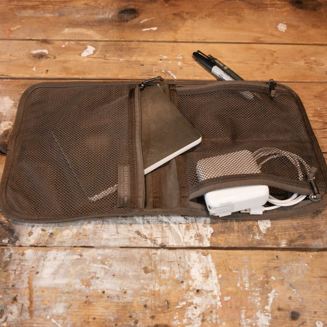 The GORUCK Wire Dopp - Waxed Canvas is a brown zippered pouch for EDC organization, holding a notebook, charger, and cables on a worn wooden surface.
