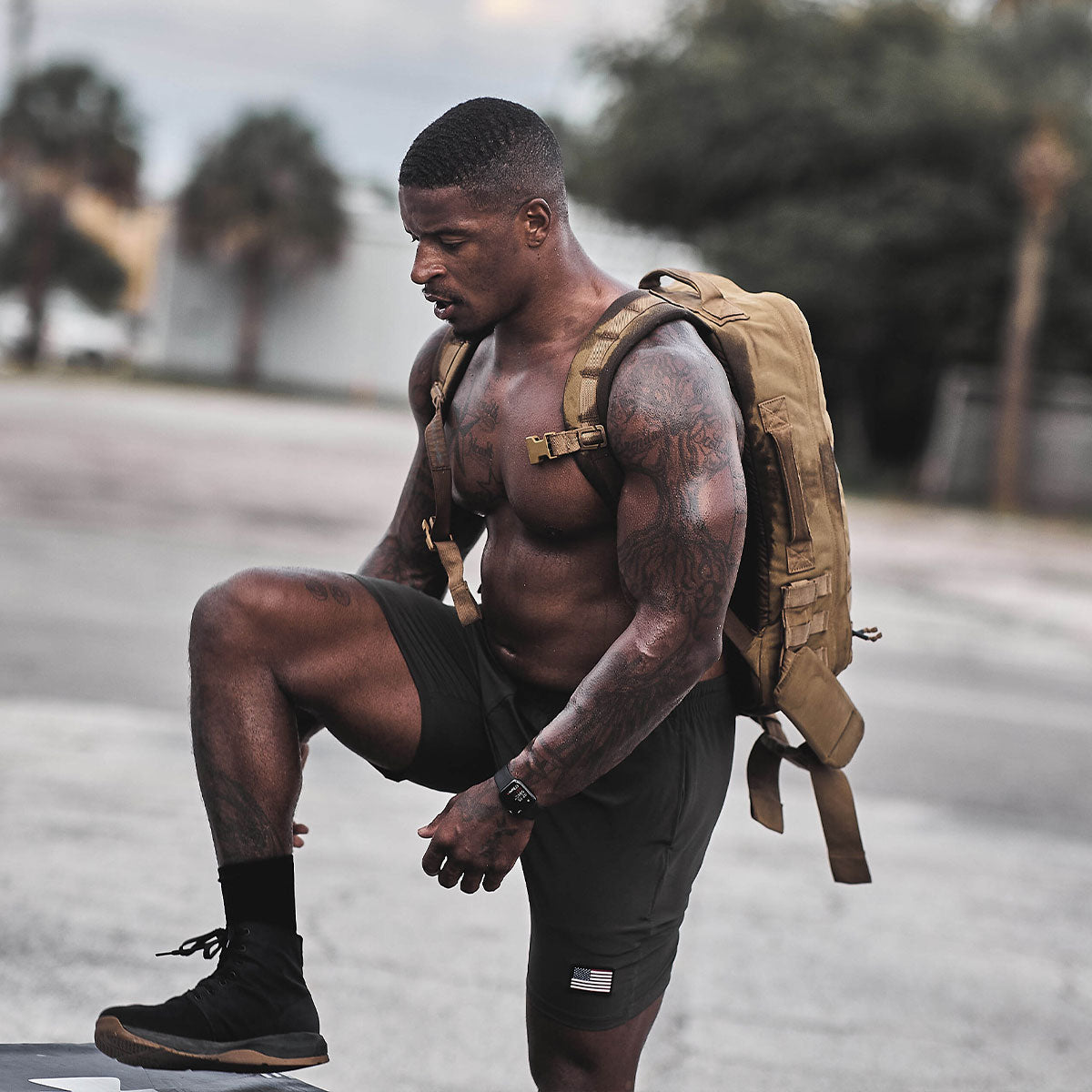 Muscular man with a backpack pauses outdoors, resting one leg on a surface, wearing black shorts and boots.