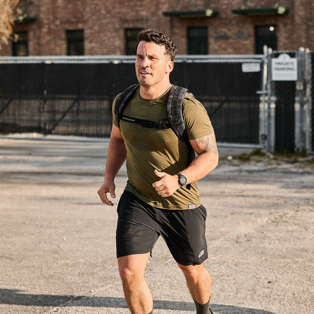 A man jogs outdoors near a fence and brick building wearing the GORUCK Ruck Plate Carrier 3.0, an athletic backpack with ergonomic lumbar support.