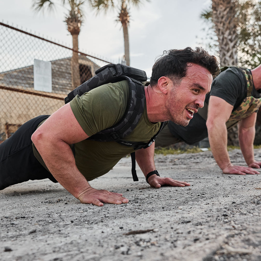 A man in workout gear uses the GORUCK Ruck Plate Carrier 3.0 to do push-ups on dirt outdoors, looking focused and determined with palm trees in the background.