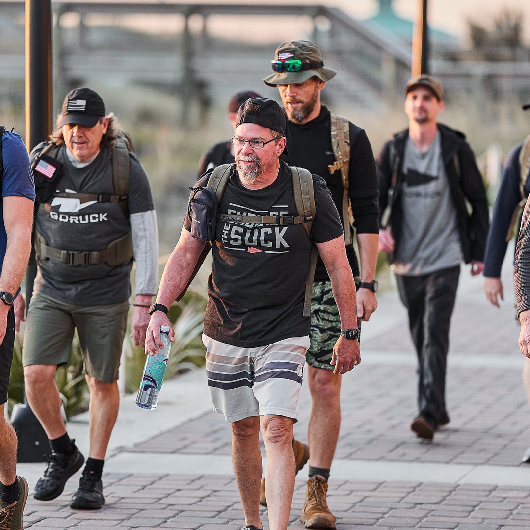 A group of men in casual athletic wear and backpacks walk outdoors, united in resilience as they wear the vendor-unknown Embrace the Suck Tee - Tri-Blend, inspired by the enduring spirit of the USMC motto.
