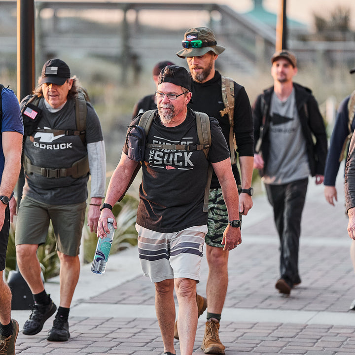 A group of men in casual athletic wear and backpacks walk outdoors, united in resilience as they wear the vendor-unknown Embrace the Suck Tee - Tri-Blend, inspired by the enduring spirit of the USMC motto.