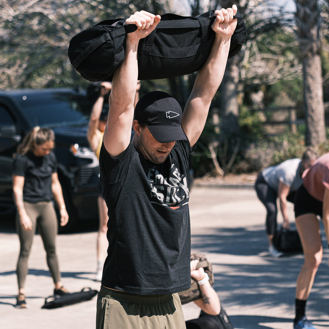 Wearing the Embrace the Suck Tee - Tri-Blend by vendor-unknown, a person lifts a heavy sandbag overhead outdoors, capturing the USMC spirit with others working out in the background.