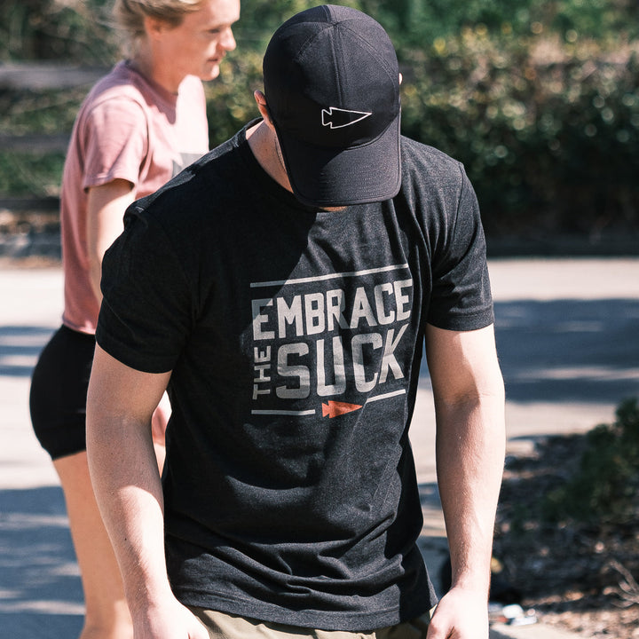 A man wearing the vendor-unknown Embrace the Suck Tee - Tri-Blend, inspired by the classic USMC motto, stands outside with another person visible behind him.