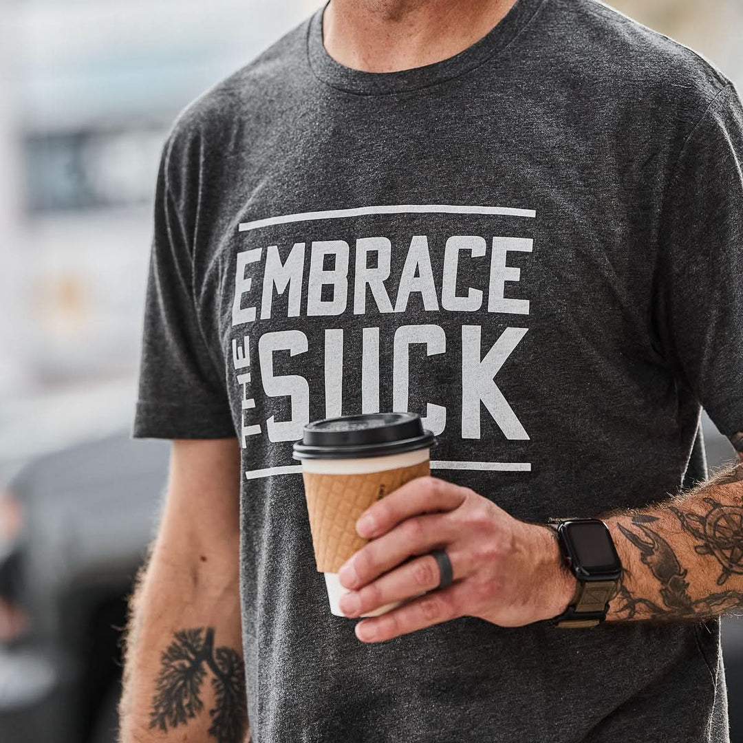 Wearing the Embrace the Suck Tee - Tri-Blend from vendor-unknown, this person holds a coffee cup and sports a smartwatch, channeling USMC resilience in daily life.