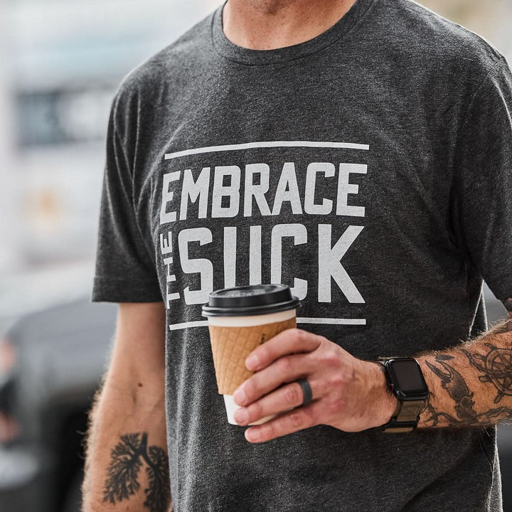 Wearing the Embrace the Suck Tee - Tri-Blend from vendor-unknown, this person holds a coffee cup and sports a smartwatch, channeling USMC resilience in daily life.