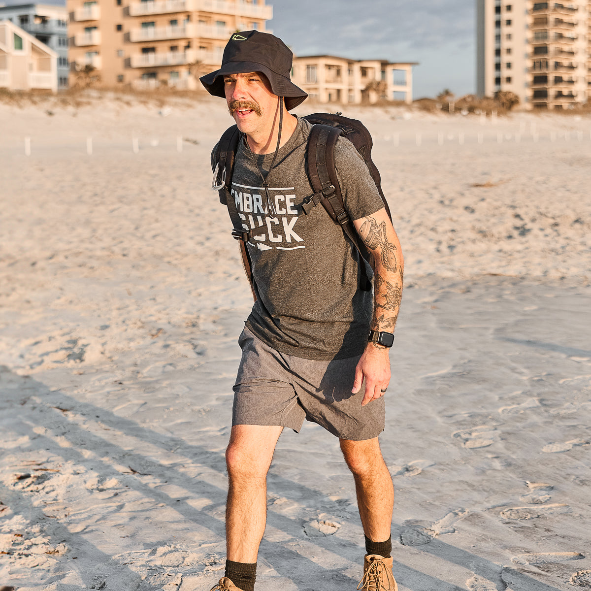 Man rucking on beach in GORUCK Embrace the Suck charcoal tee, shorts, and backpack.