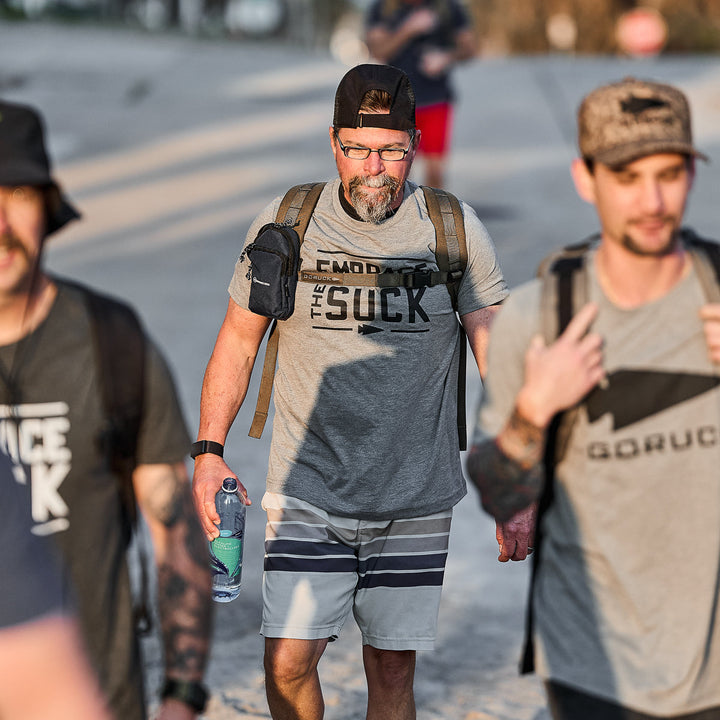 Three men in vendor-unknown’s Embrace the Suck Tee - Tri-Blend walk outdoors, one with a backpack and water bottle. All look focused, embodying determination and resilience through the USMC-inspired motto.