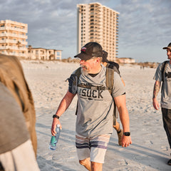 Man wearing GORUCK Embrace the Suck tee and backpack walking on beach with water bottle
