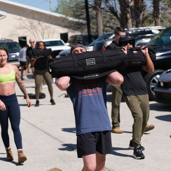 Outdoor group fitness with weighted sandbags—embrace the challenge in style with the vendor-unknown Embrace the Suck Tee - Tri-Blend, inspired by the official USMC motto.