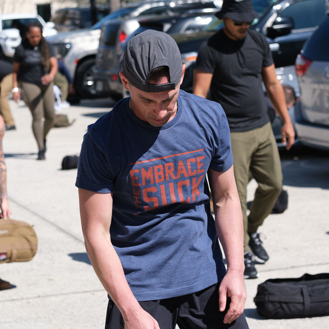 A man wearing the vendor-unknown Embrace the Suck Tee - Tri-Blend, inspired by an official USMC motto, and a backwards cap stands outdoors with others and exercise gear nearby.