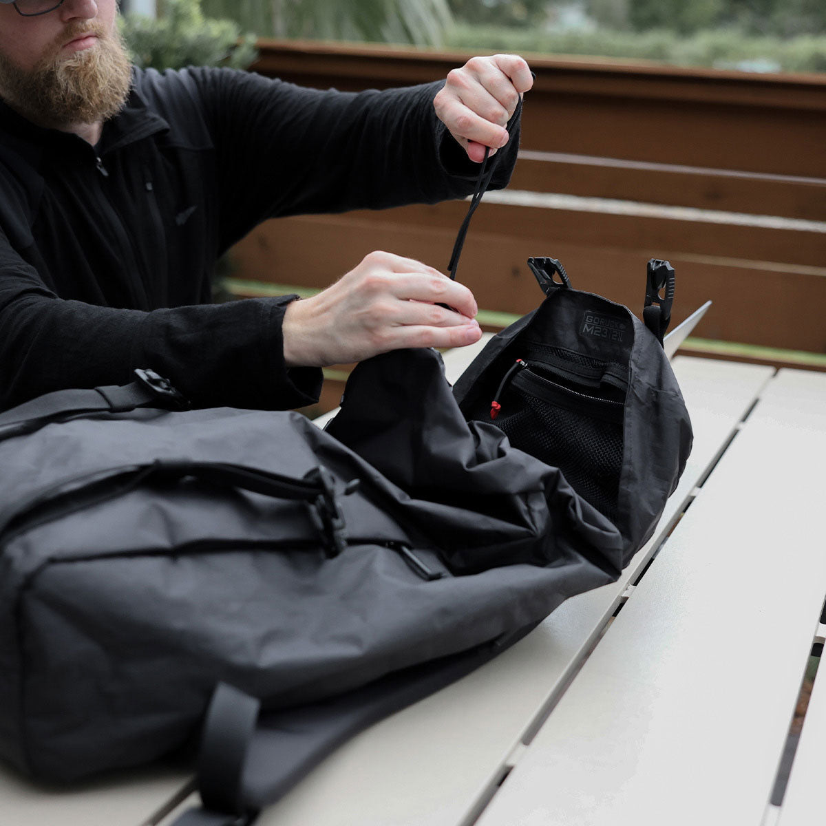A person unzips a black backpack on a light-colored outdoor table.