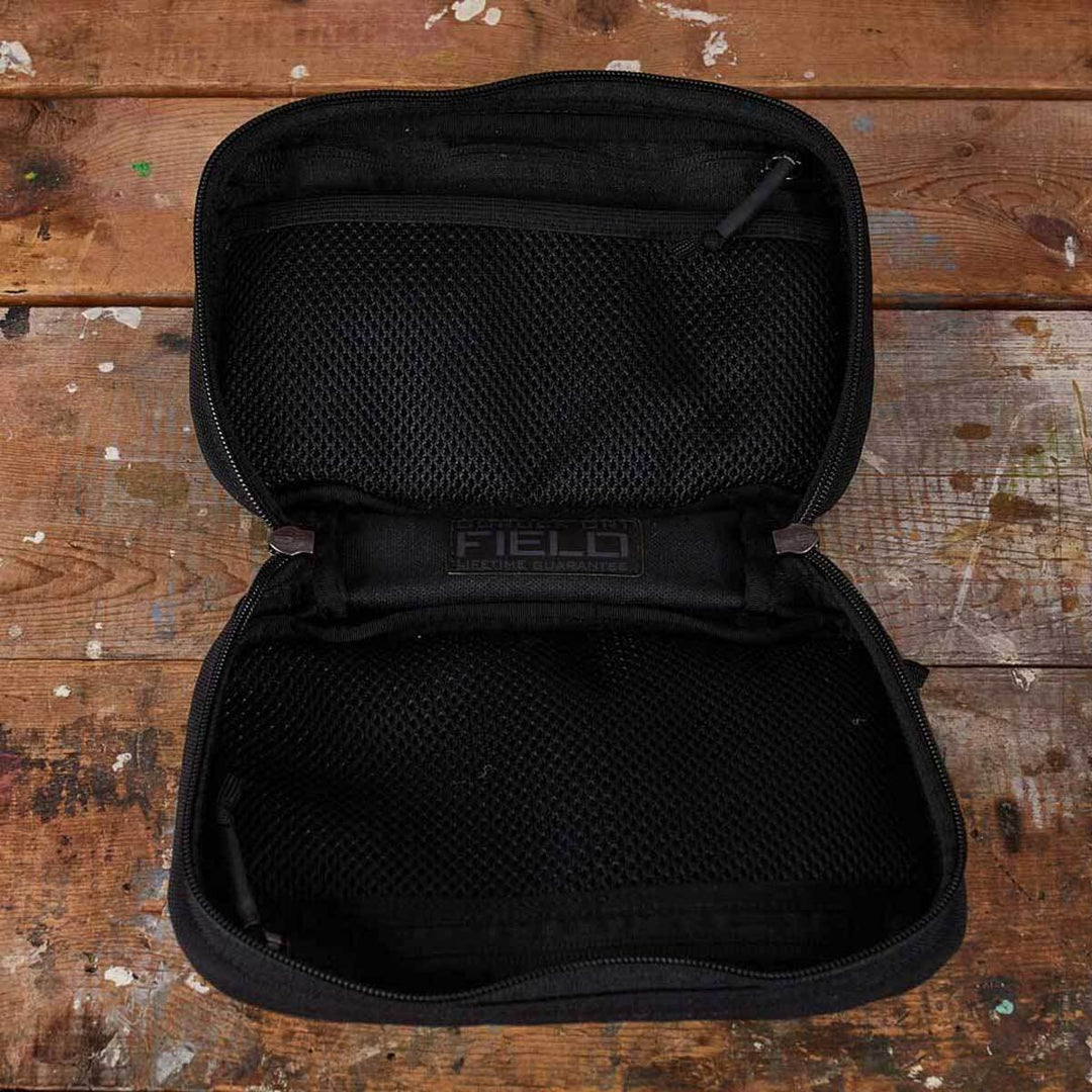 The GR1 Field Pocket by GORUCK, a black travel case with mesh pockets ideal for organizing camera gear, rests on a rustic wooden surface.