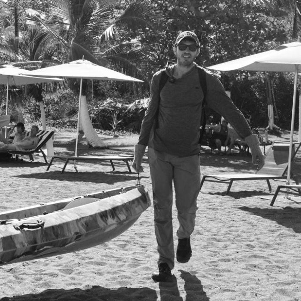 A man in sunglasses carries a kayak on a sandy beach with umbrellas and palm trees in the background.