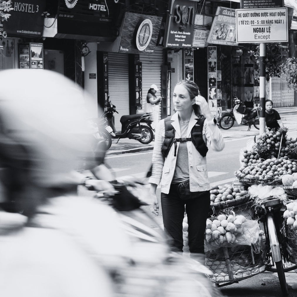 A woman stands near fruit carts on a busy street, as blurred motorbikes pass by.