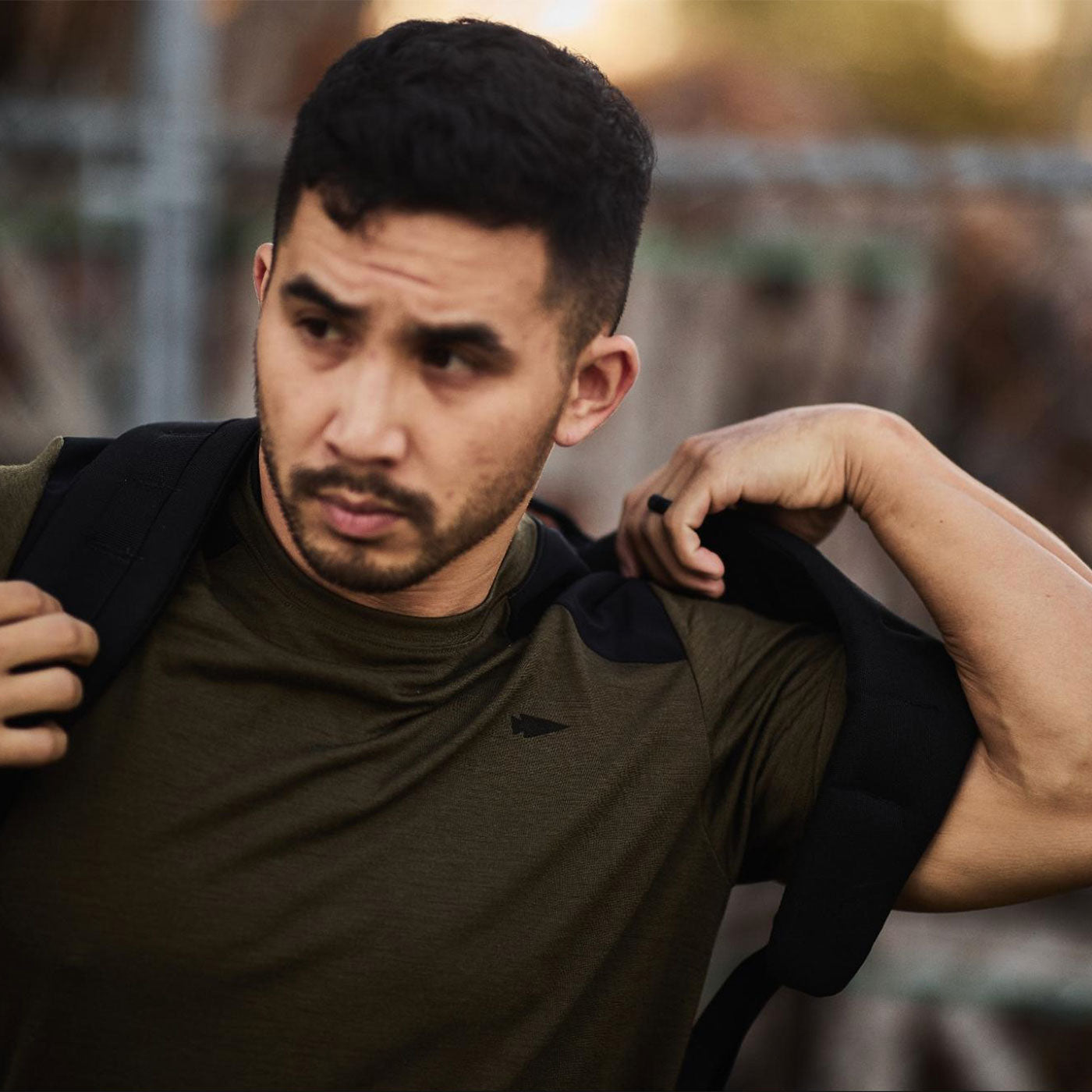 Man in an olive green shirt puts on a black backpack outdoors, looking to the side with a serious expression.