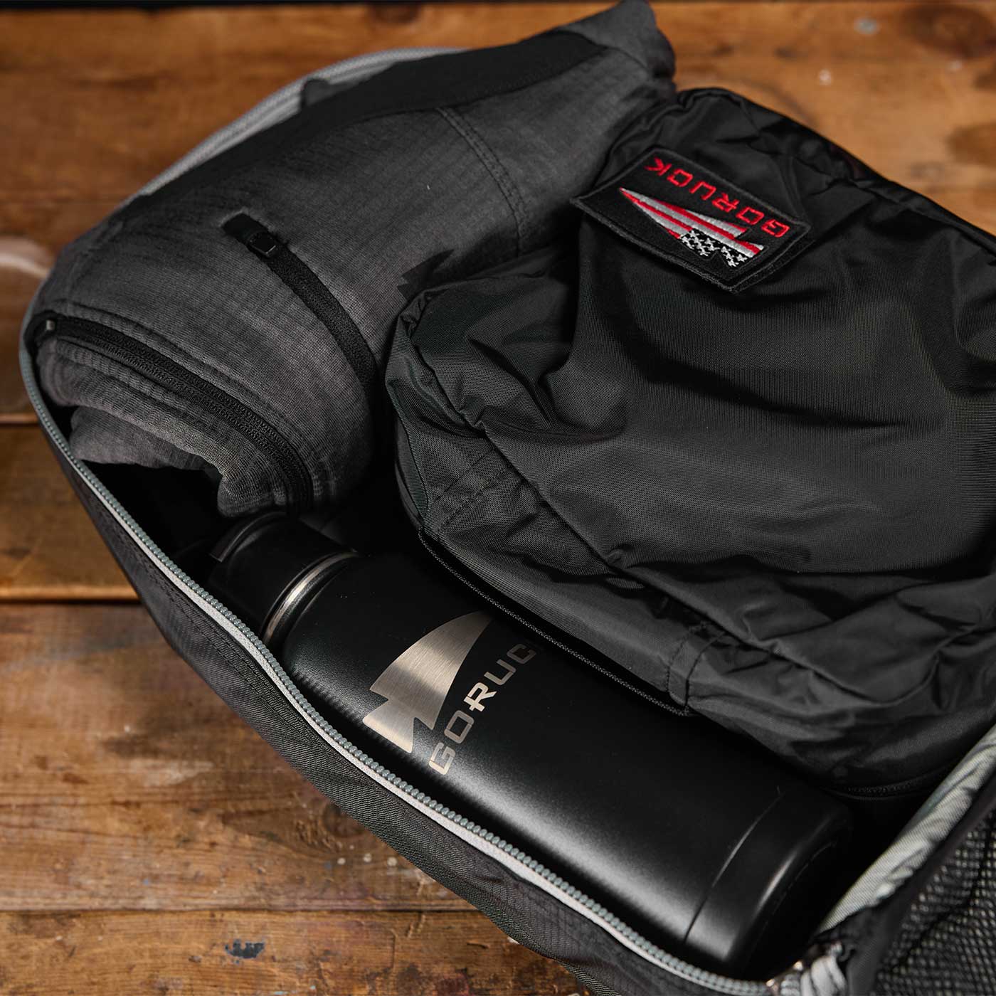 Open backpack with a water bottle, folded clothing, and a black GORUCK pouch on a wooden surface.