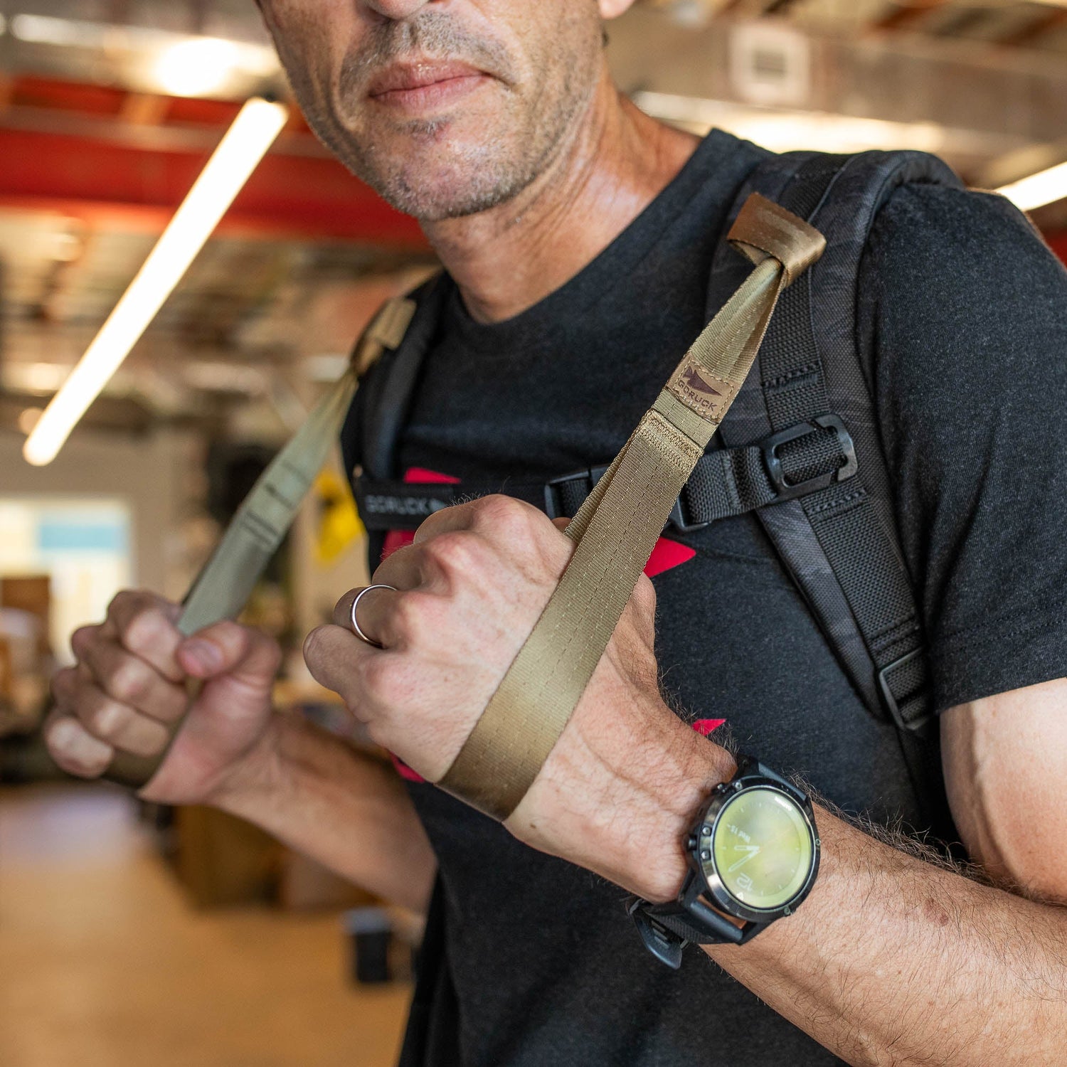 A man grips the T-REX Hand Straps, attached to his backpack straps, while wearing a black t-shirt indoors.