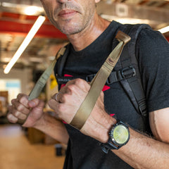 A man grips the T-REX Hand Straps, attached to his backpack straps, while wearing a black t-shirt indoors.