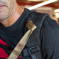 Close-up of a man wearing a black shirt and black backpack with tan T-REX Hand Straps over his shoulder, featuring an anchor loop for attaching gear or a med band.
