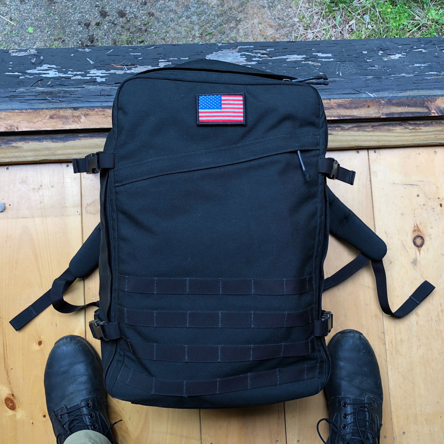 A black backpack with a U.S. flag patch on top, placed on a wooden floor next to black boots.