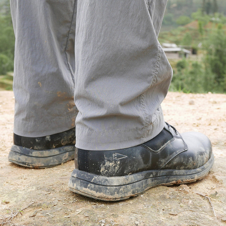 Person wearing muddy black shoes and gray pants standing outdoors on a dirt surface.