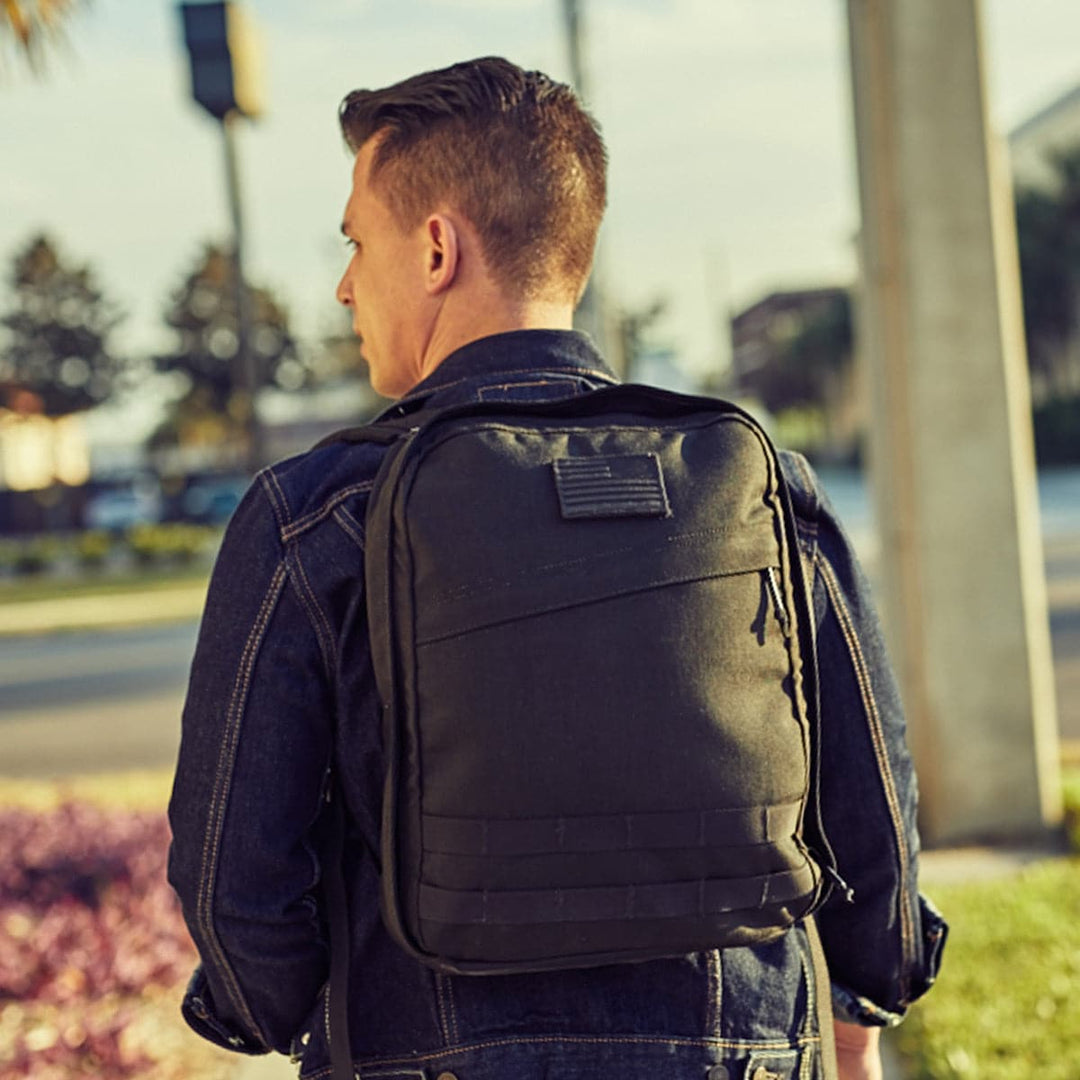 A person wearing a denim jacket is seen from behind, walking outside with a compact GORUCK GR0 - Cordura backpack. The bag, featuring a bombproof laptop compartment, stands out against the blurred urban backdrop of trees and structures under a clear sky.