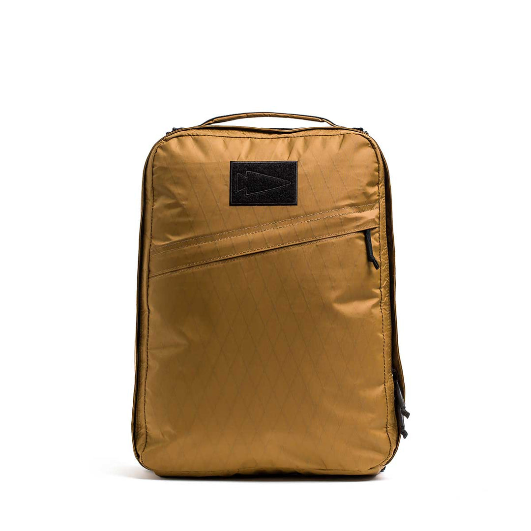 The GORUCK GR0 - X-PAC is a gold rectangular backpack made from lightweight X-PAC material, featuring a diagonal zippered pocket and a black front patch, shown against a white background.