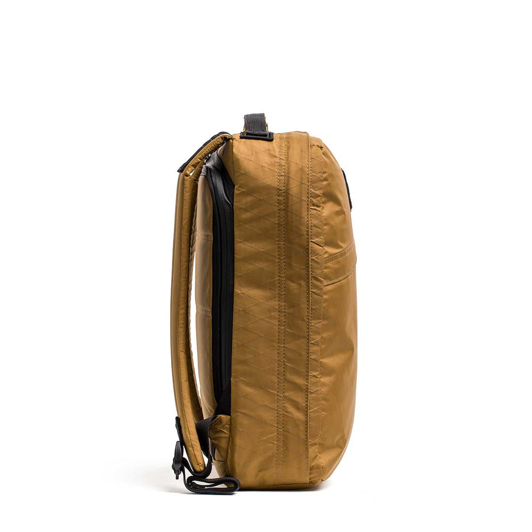 Side view of the GORUCK GR0 - X-PAC backpack in brown, featuring padded straps and a zippered compartment. This slim, lightweight design delivers modern style and durability while standing upright.