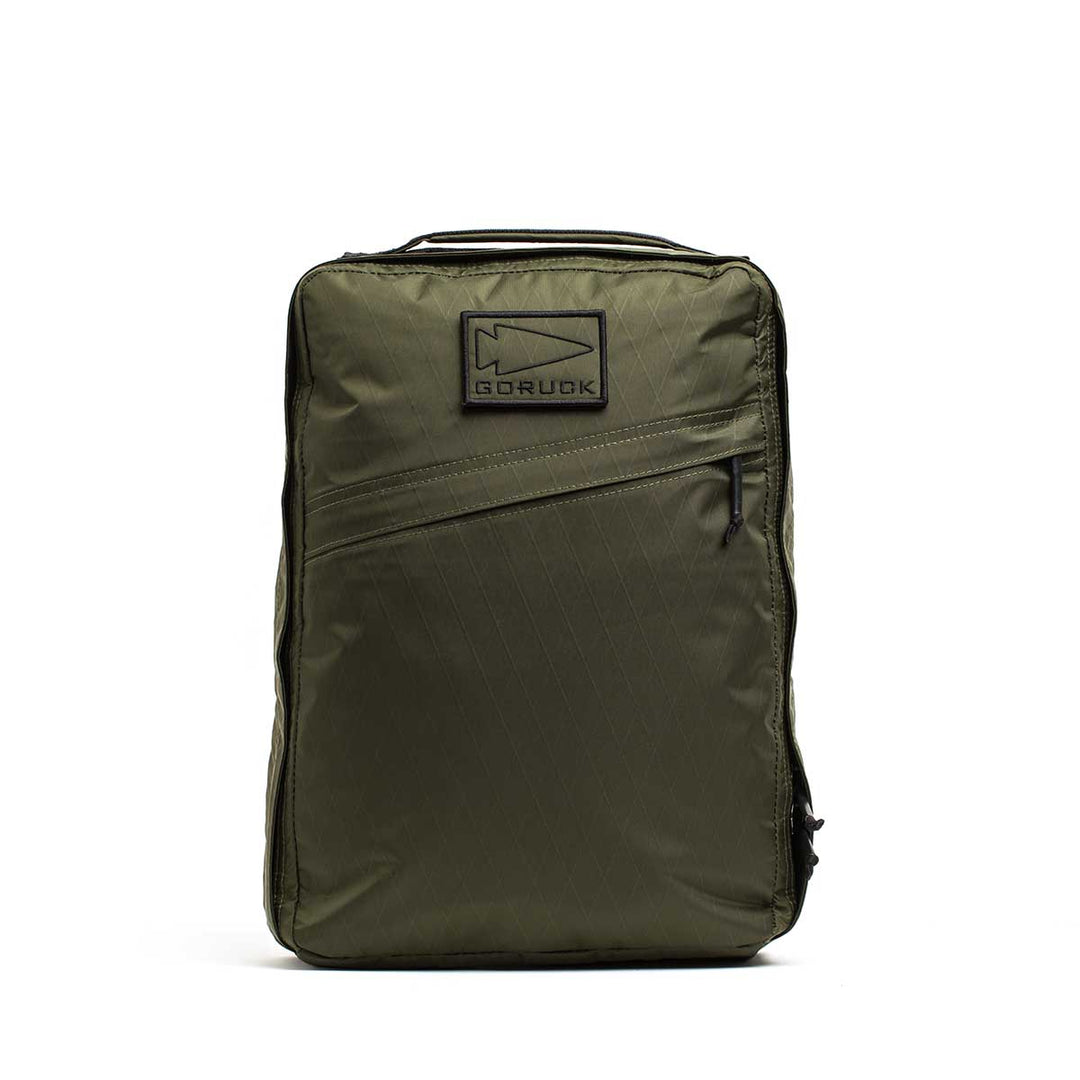 The GORUCK GR0 - X-PAC is an olive green, lightweight backpack with a front zipper pocket and a rectangular logo patch on the upper front, designed for everyday use.