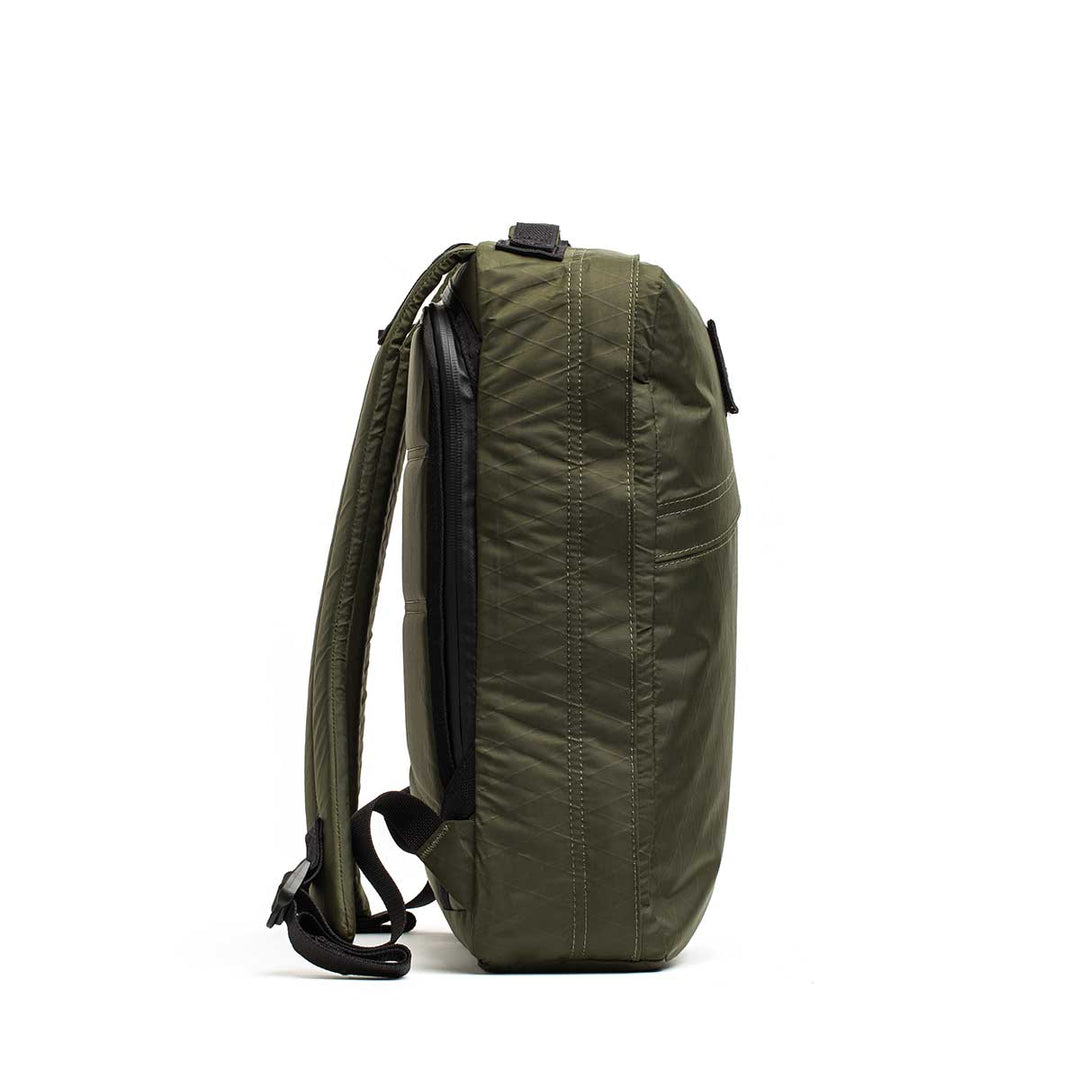 A side view of the olive green GORUCK GR0 - X-PAC backpack with black straps and zipper, standing upright on a white background.