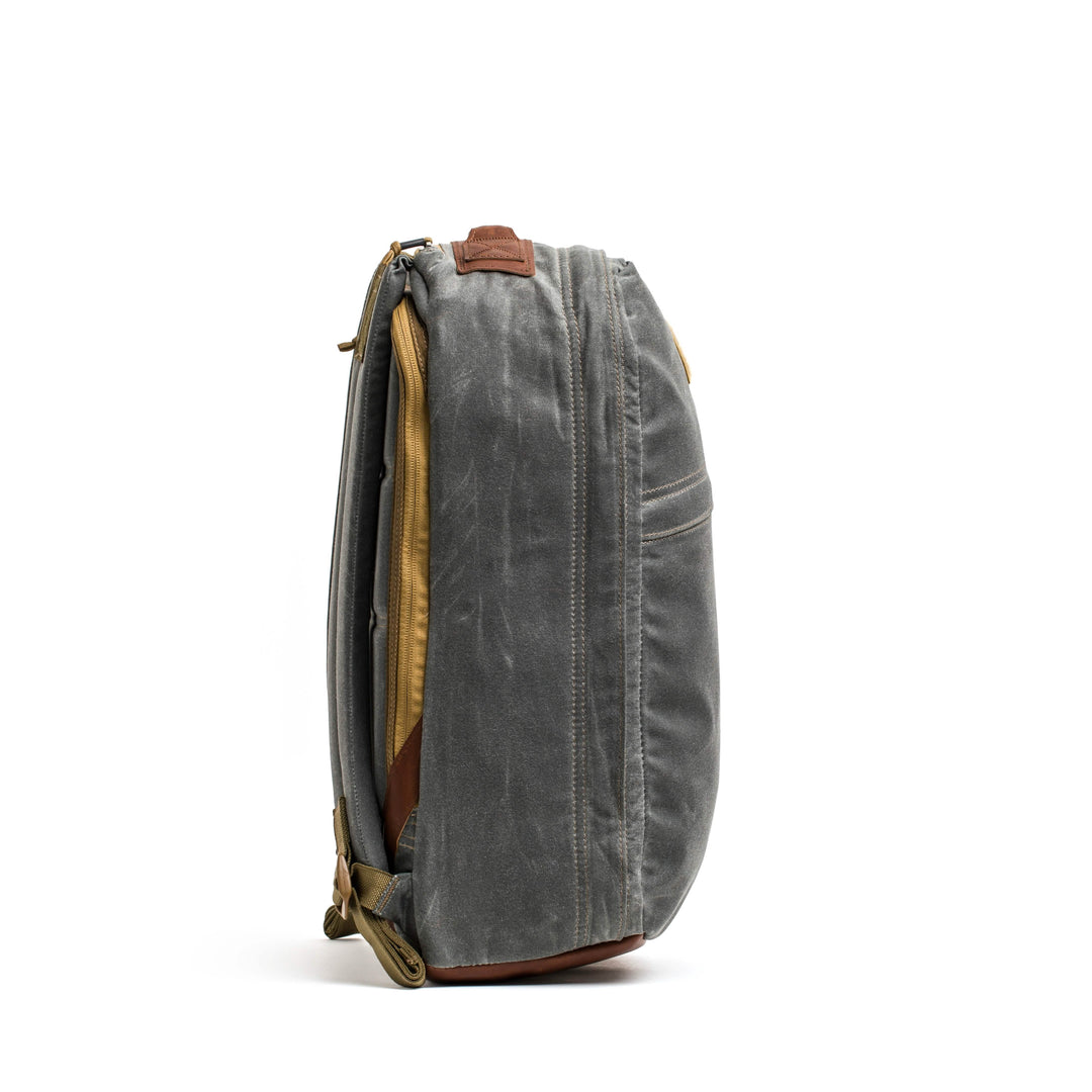 The GORUCK GR1 USA - Heritage backpack features a side view of gray waxed duck canvas with brown leather accents, crafted from durable Martexin Wax Finish. It has retro-inspired design elements and multiple zippered compartments for versatile storage.