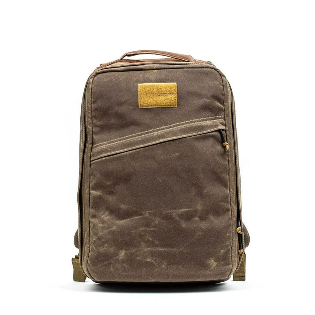 The GORUCK GR1 USA - Heritage is a brown rectangular rucksack made from waxed duck canvas, featuring a front zipper pocket and a Red Wing leather patch on top.