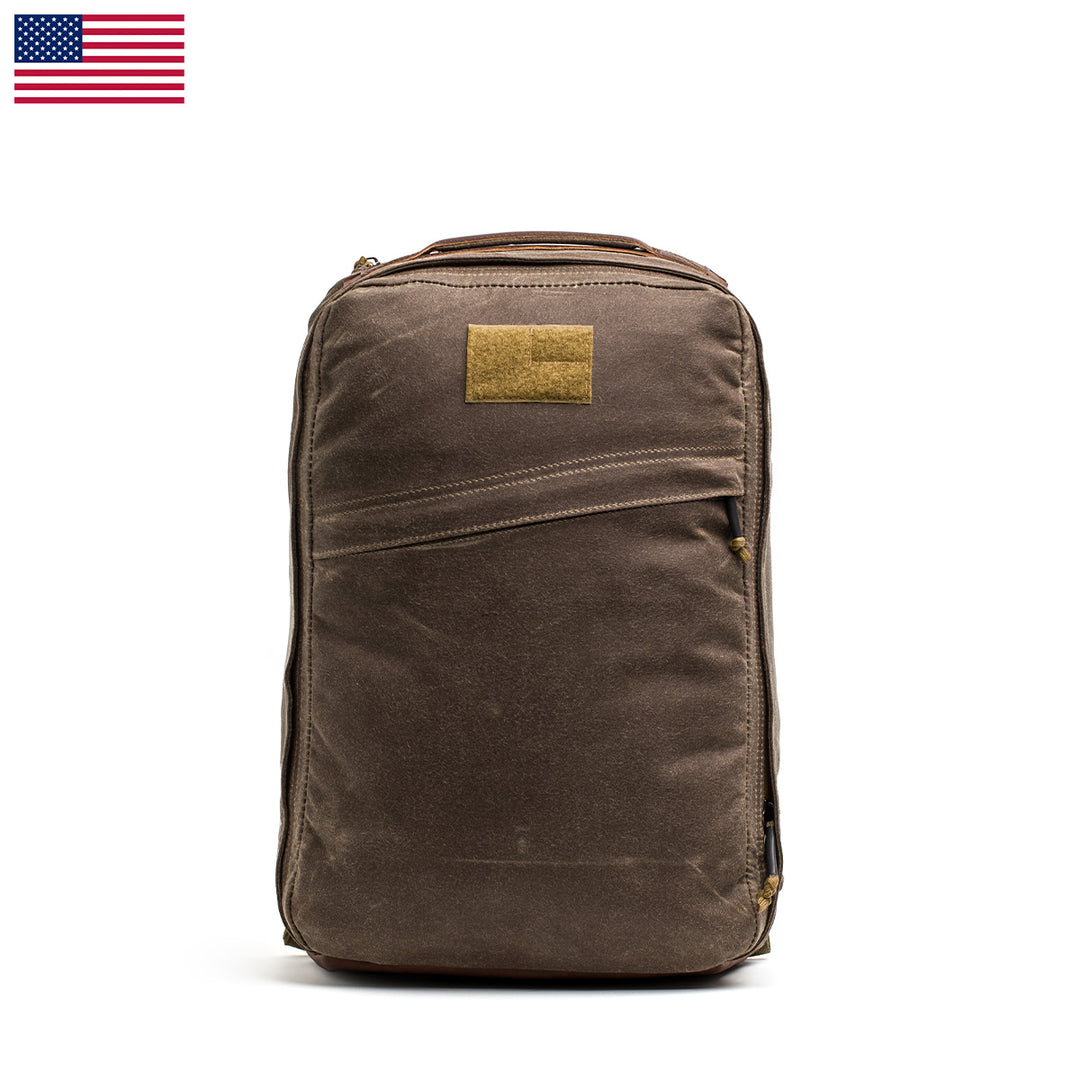 The GORUCK GR1 USA - Heritage backpack, featuring a brown design with a small tan patch, stands upright on a white background with the U.S. flag in the top left corner.